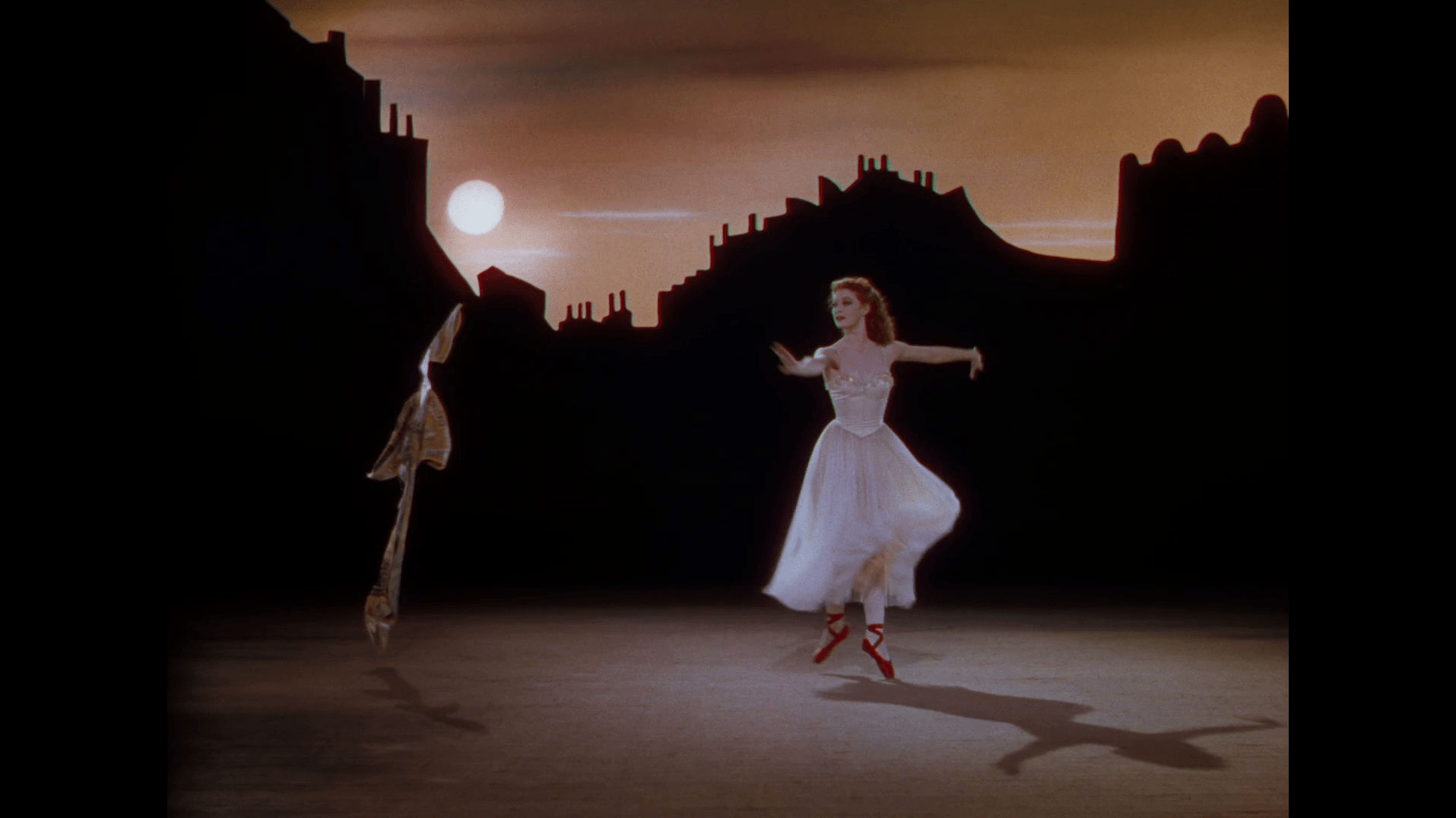 A white woman with red hair in a 40s style white ballet costume and red pointe shoes dancing with arms outstretched across a scene of a cityscape at dusk. 