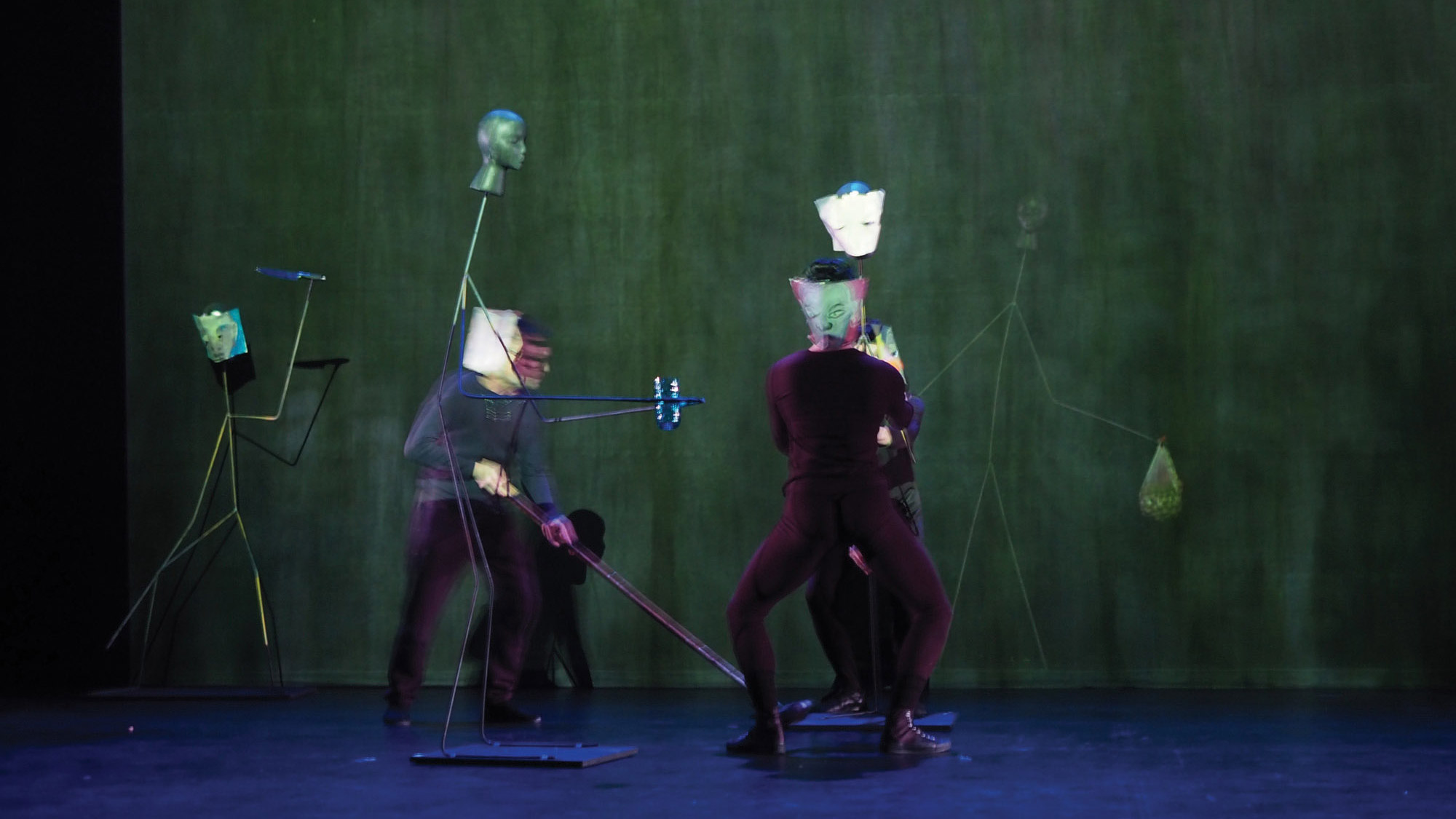 Two people preforming with various humanoid sculptures on a blue and green stage. 