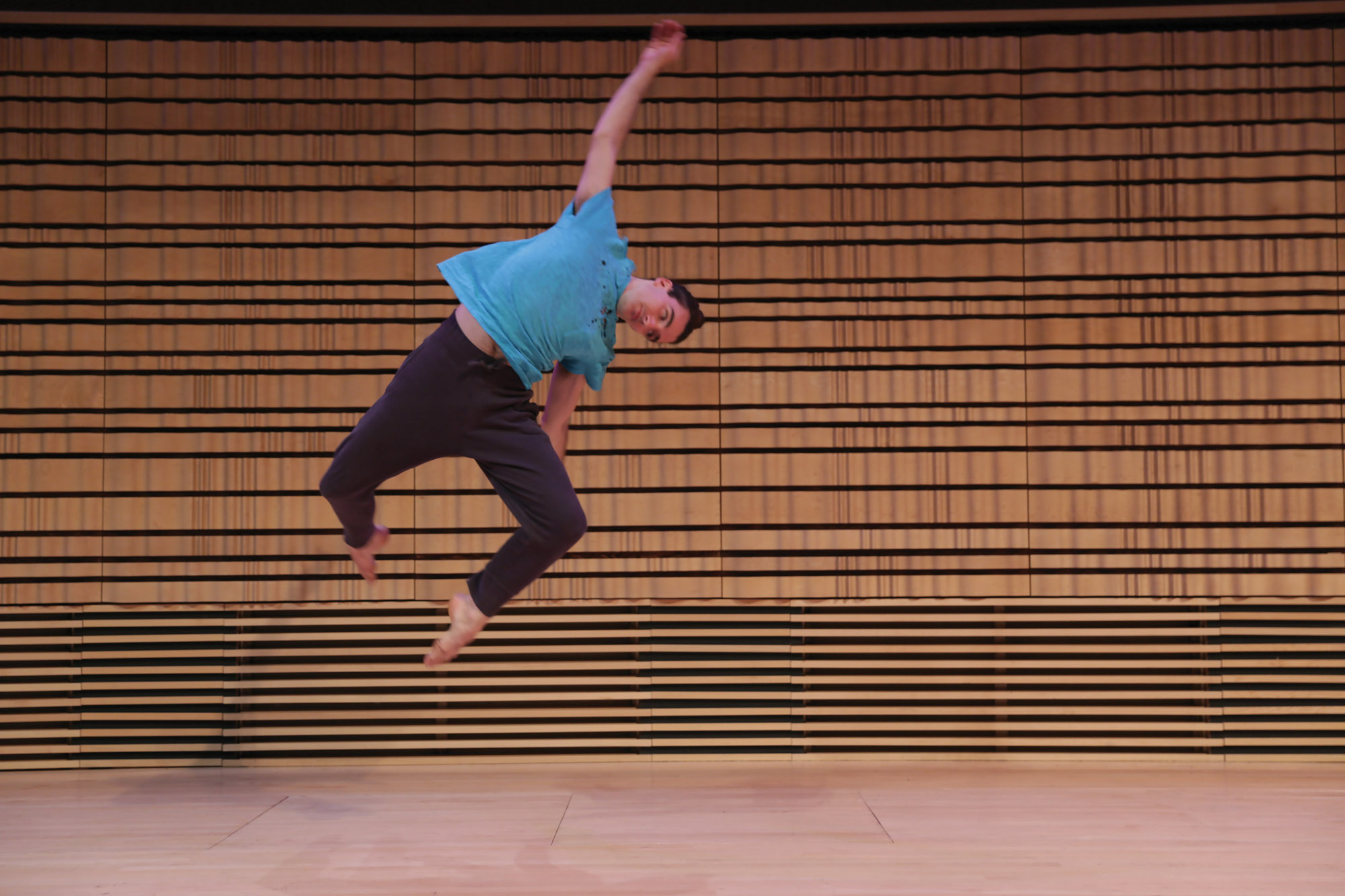 a male dancer wearing a teal top and gray bottoms suspended mid-air after a jump on the concert hall stage. 