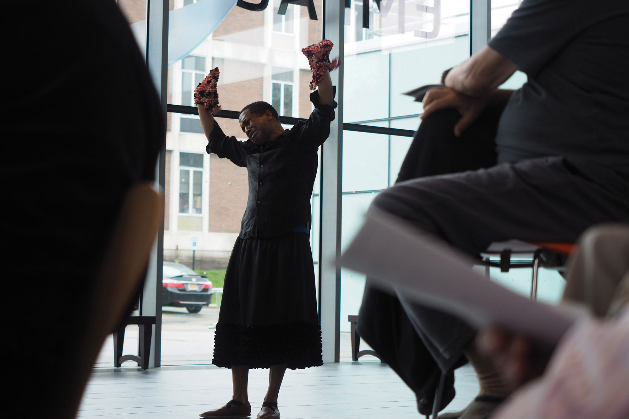 Trajal Harrell wearing a black dress performing with hands up in a light filled lobby. 