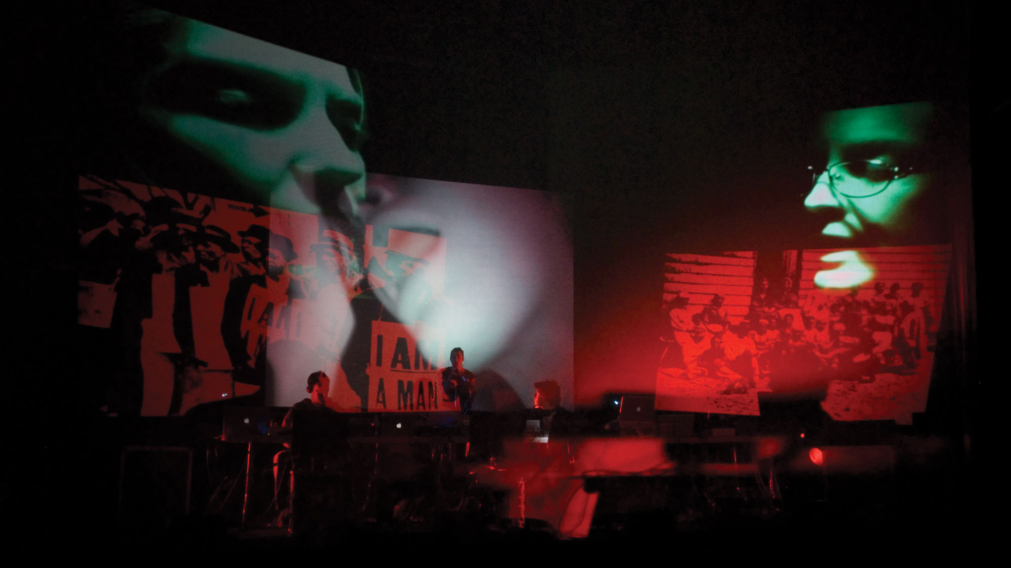 Three faces in green projected onto a dark stage with red images of protesting overlapped. A small pit orchestra is in front of them. 