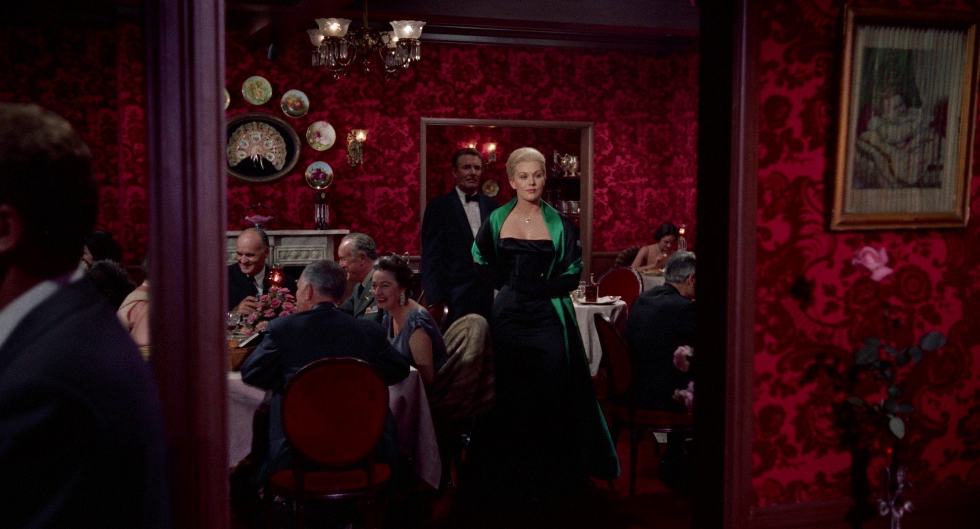 Kim Novak wearing a green 1950's cocktail dress entering a crowded restaurant with red damask wallpaper. 
