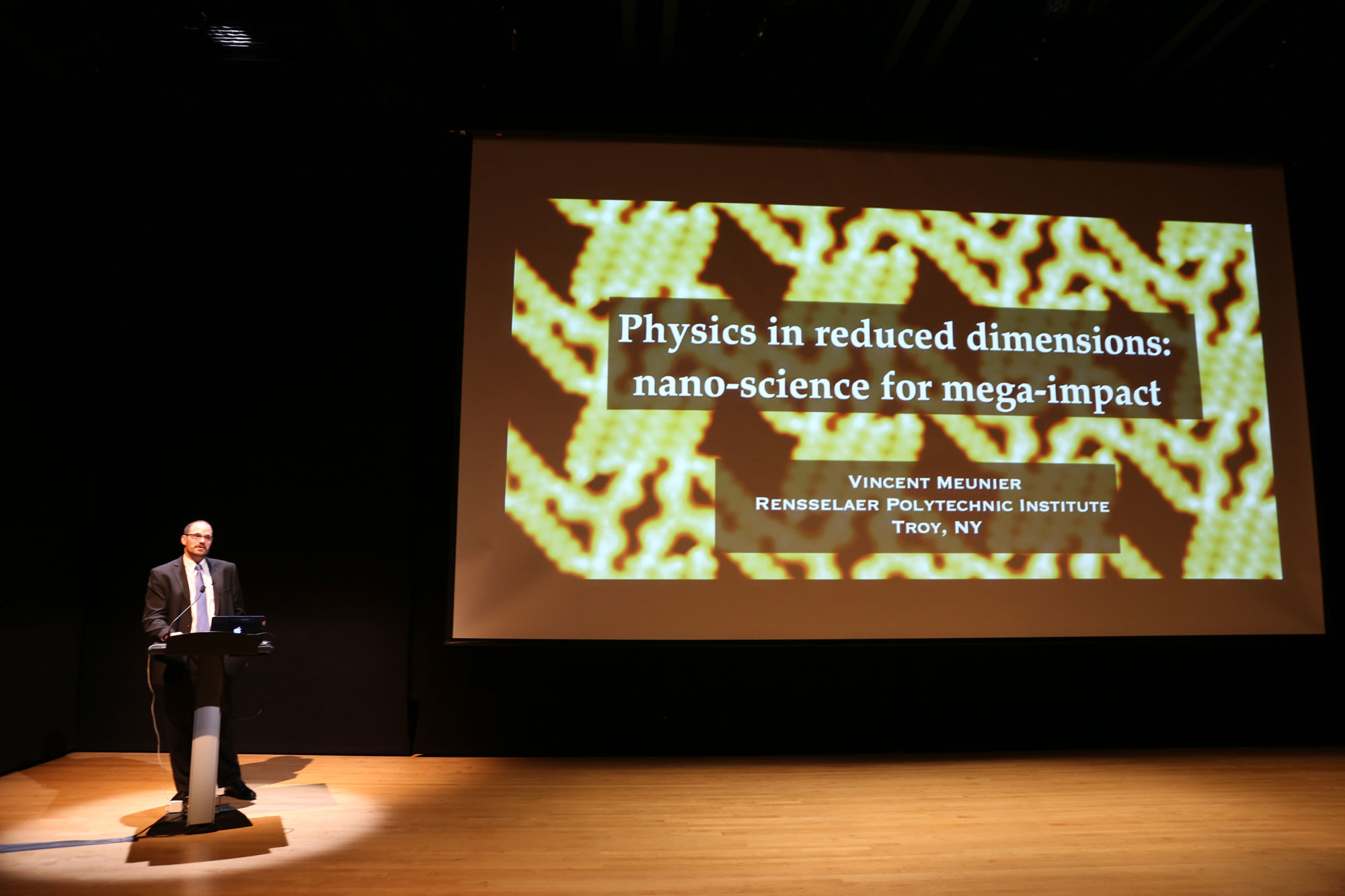 Vincent Meunier in discussion on a stage in front of a projection that reads 'Physics in Reduced Dimensions: Nano-Science for Mega-Impact'. 