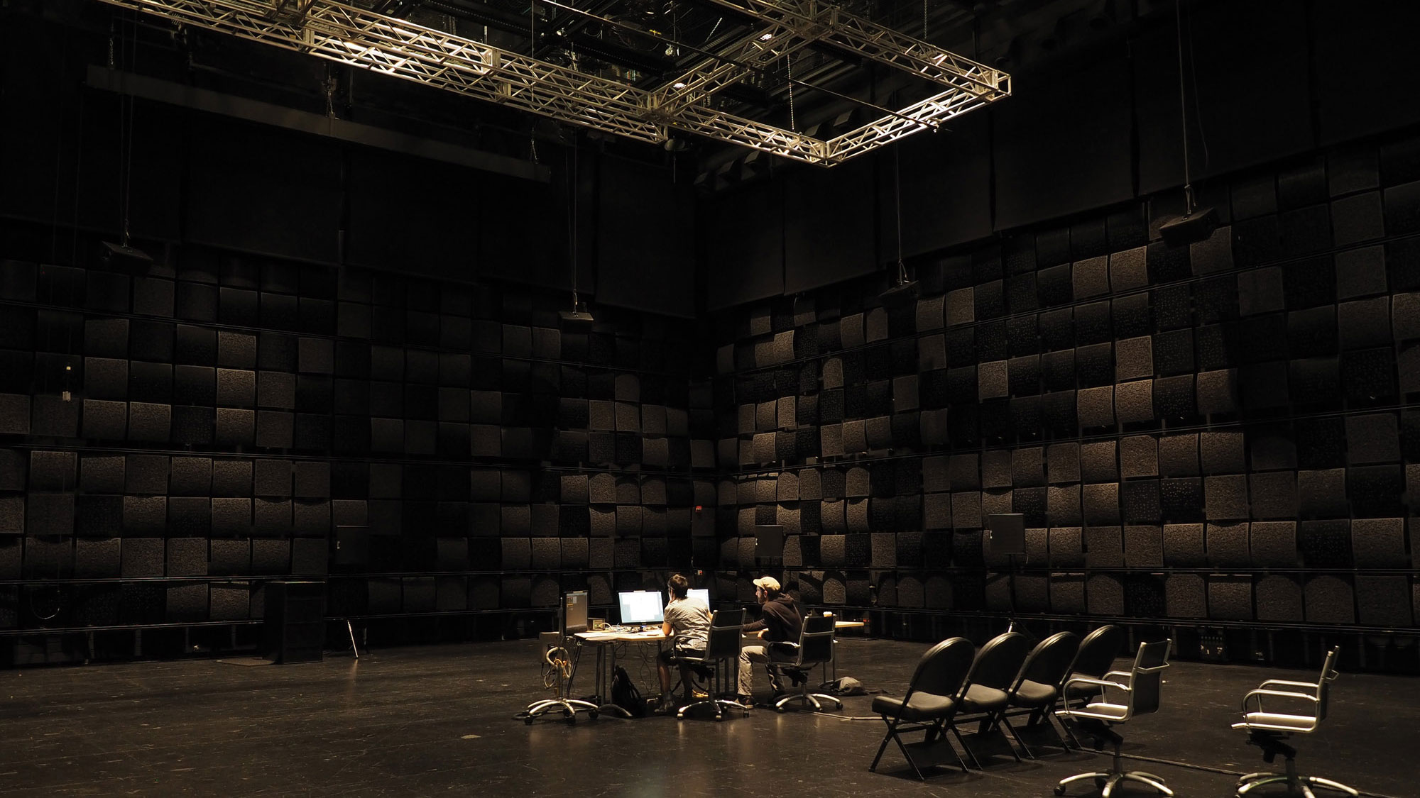 A black box studio with black acoustic tiles on the wall. Two people sit in the middle of a room at a desk with two computers below a large rectangular grid suspended from he ceiling. 