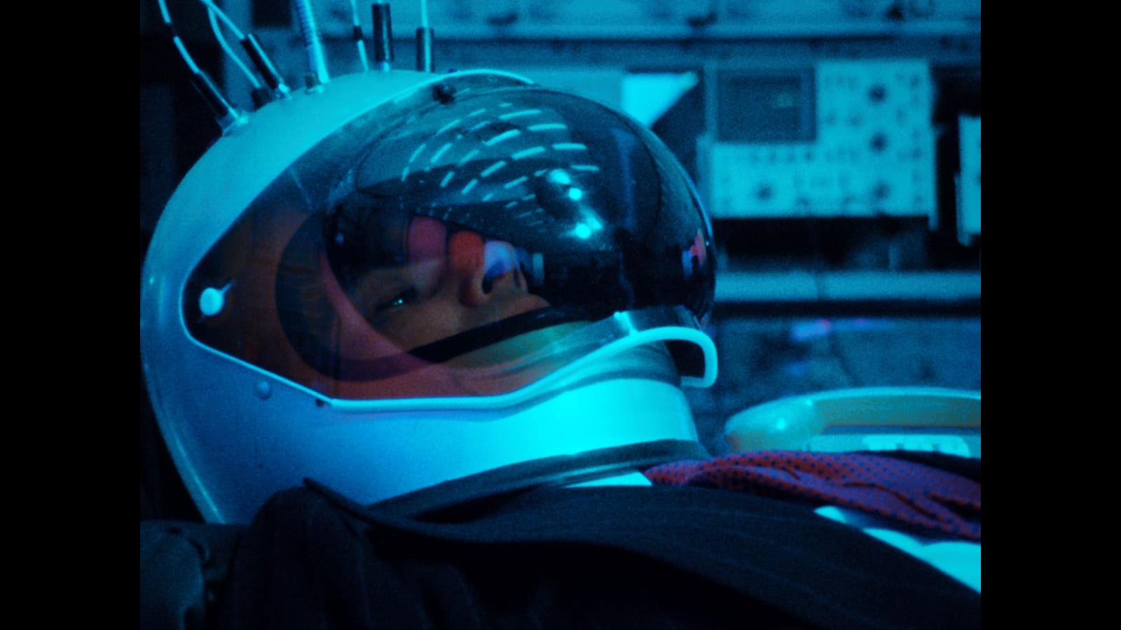 A man in a suit wearing an astronaut helmet reclined in room with various nobs and buttons washed in teal light. 