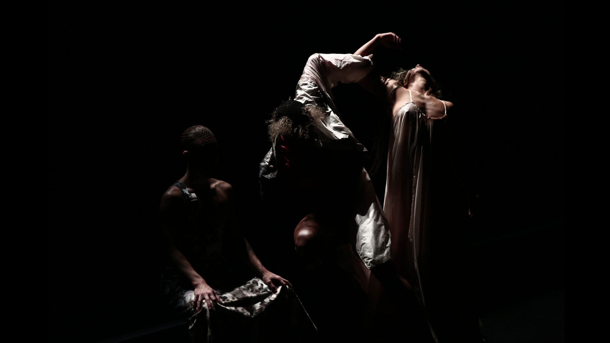 Three performers in chiaroscuro lighting. One figure is seated, another standing with arm intertwined with the third dancer who arches their back dramatically white standing. 