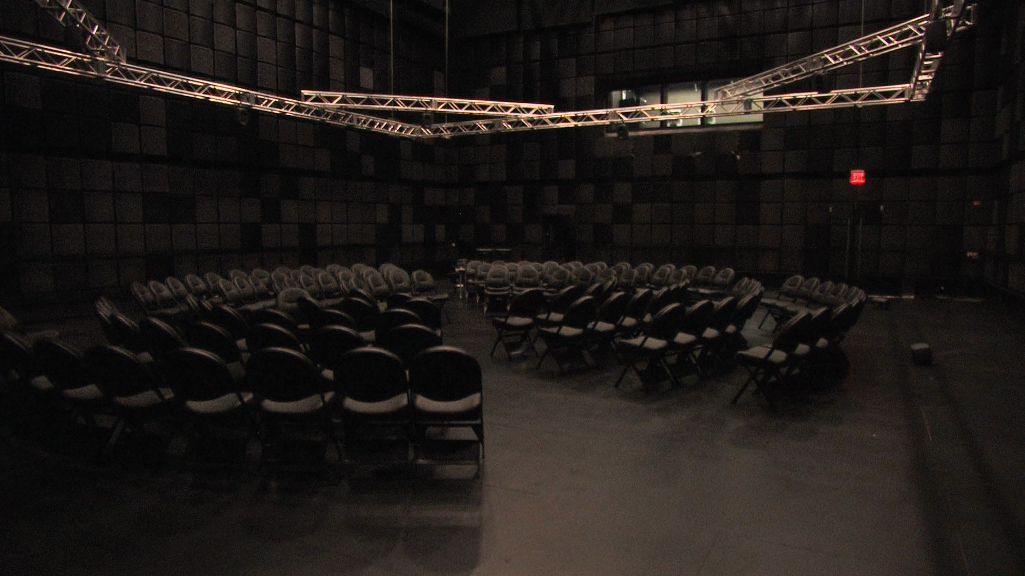 Many black folding chairs arranged in circle in a black box studio under a system of metal rigging suspended from the ceiling. 
