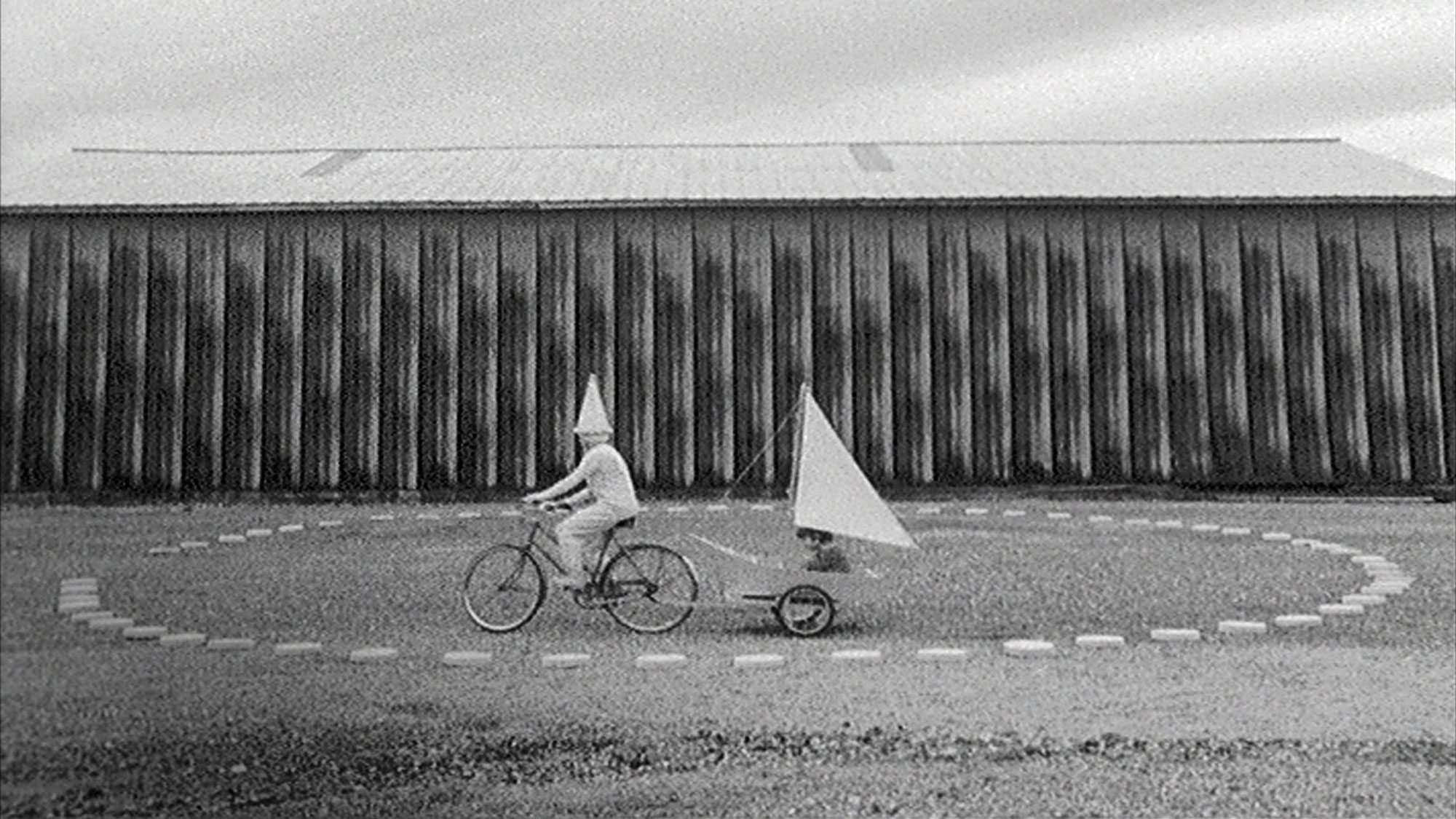  Person wearing a white cone on there head riding a bike pulling a sailboat in a circle of pavers in front of a barn. 