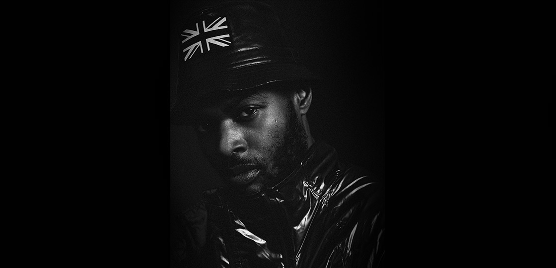 A Black man wearing a leather bucket hat with he union jack on the front and a patent leather coat looking directly at the camera in dramatic lighting. 