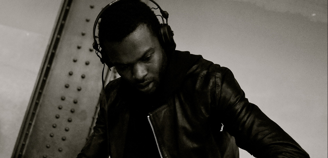 A black man wearing a leather jacket and over the ear headphones looking down with arms outstretched, focused on something of screen. 