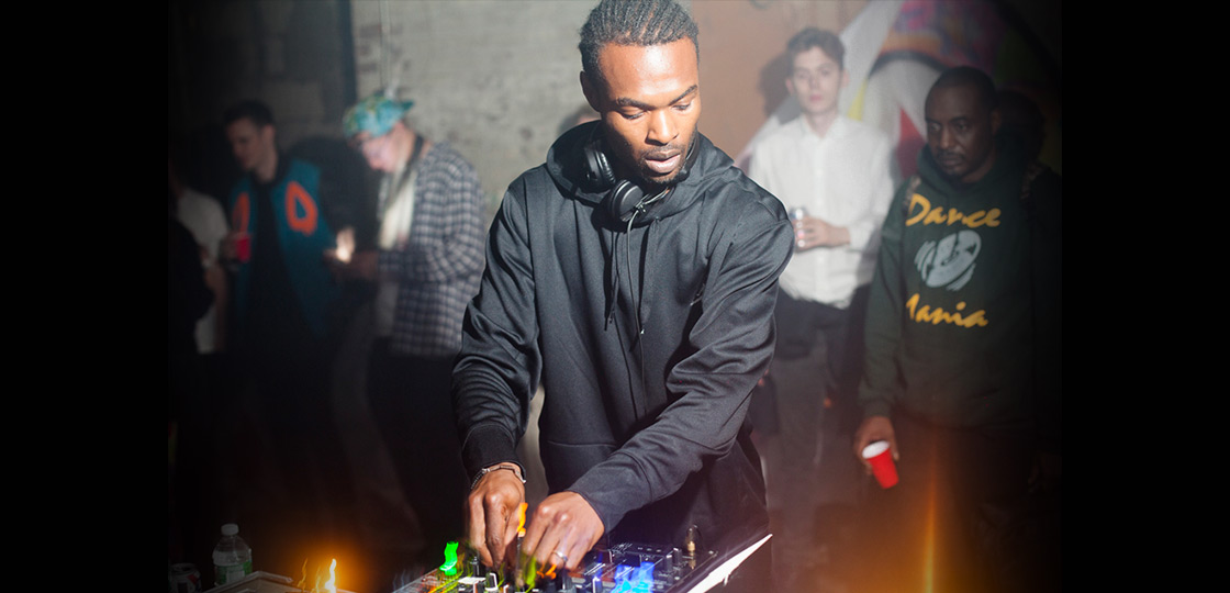 A black man wearing a black hoodie and headphones around his neck looking down focusing on a sound board and DJ equipment in front of him. A small crowd is behind him leaning against a concrete wall. 
