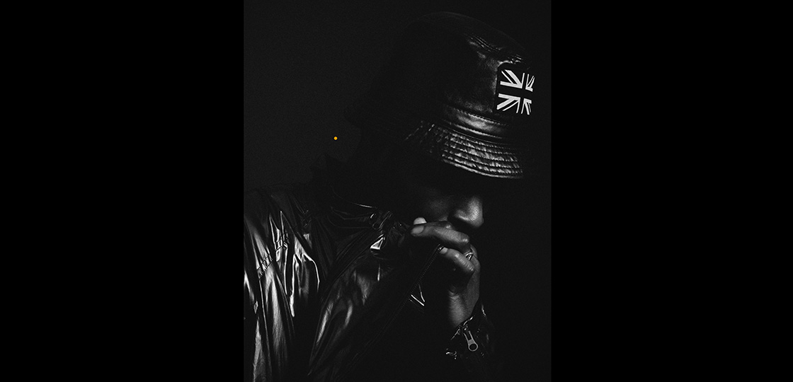 A Black man wearing a leather bucket hat with he union jack on the front and a patent leather coat looking down with his hand over his mouth. 