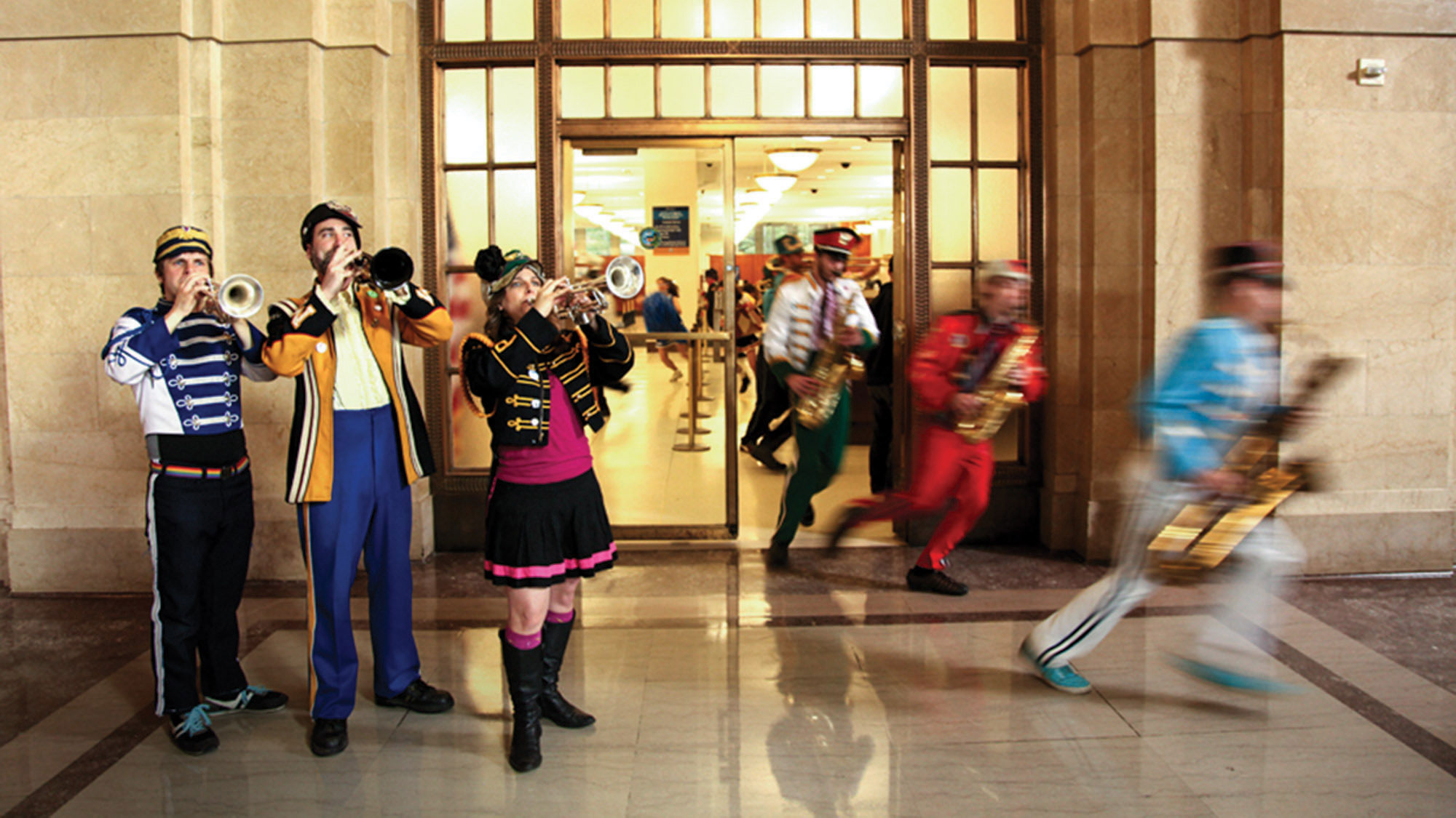 A marching band wearing traditional marching band costumes in various colors running out the door of an office or government building. 