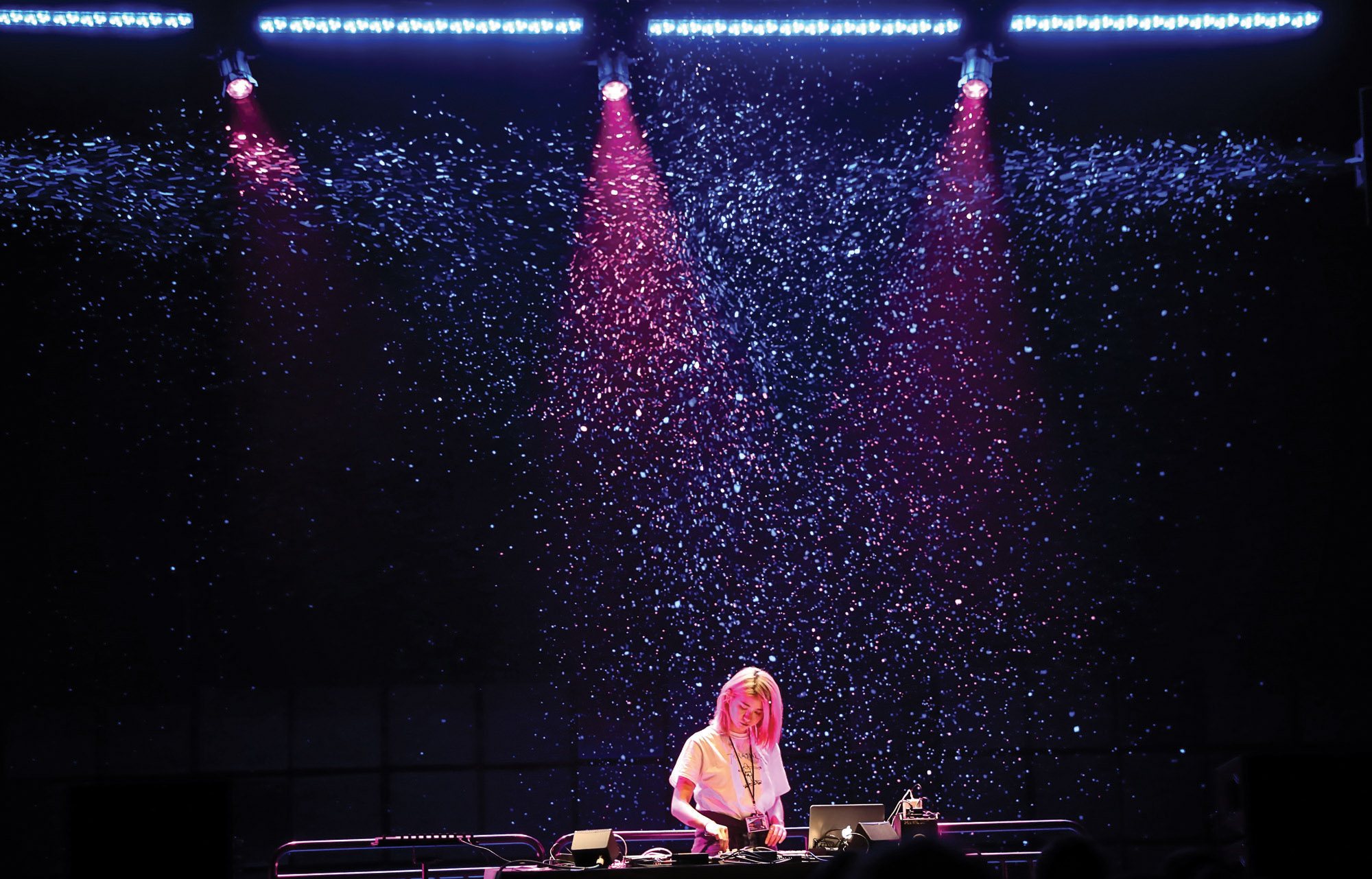 Qrion DJing on stage in front of a wall of pink and blue star-esque lights. 