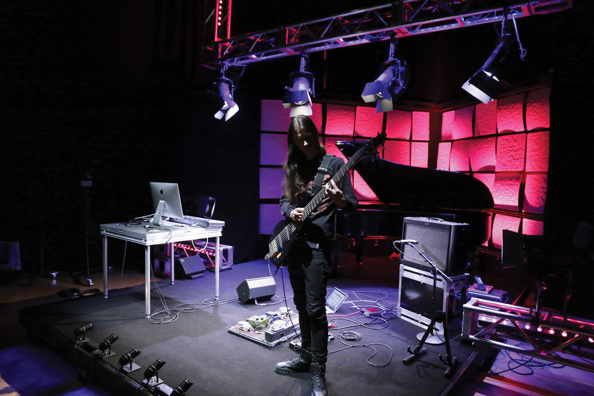 Colin Marston playing bass on a stage cluttered with amps washed in red and blue light. 