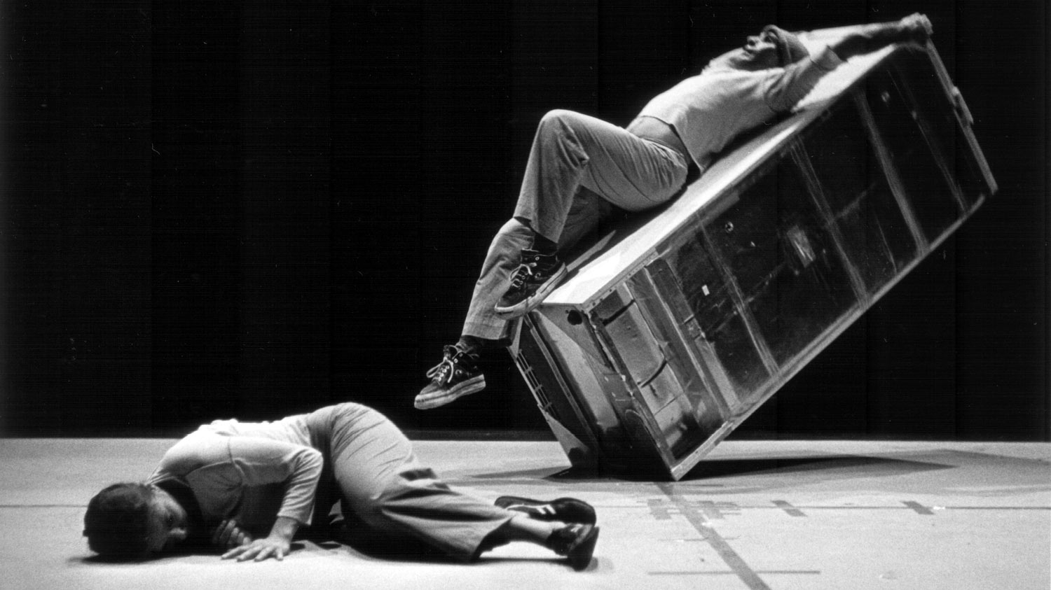 A woman lying upstage of a man laying across a refrigerator at a 45 degree angle about to tip over. 