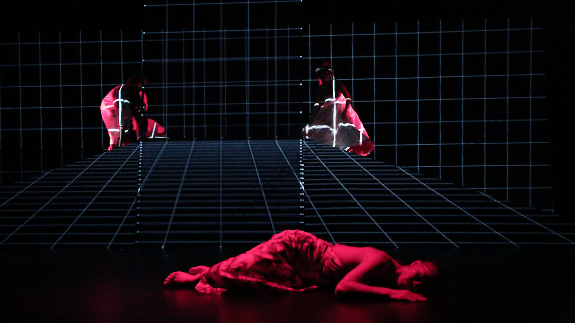 A person laying on a black stage draped in fabric and washed in red light in front of a blue grid. 
