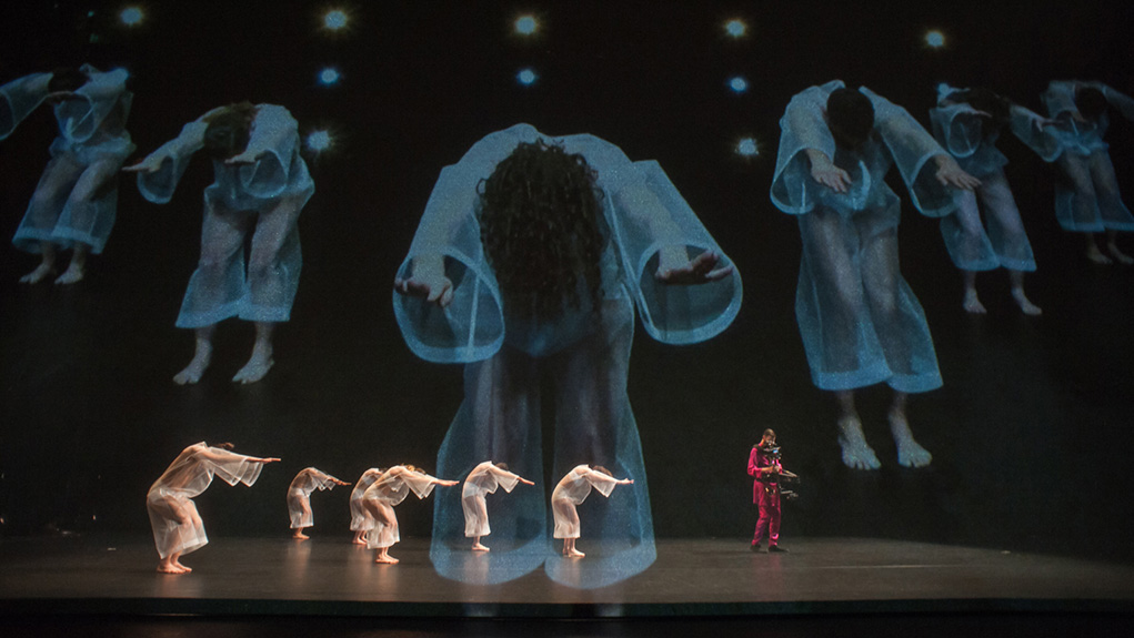 Six dancers dressed in sheer kaftans on stage with a stedicam operator wearing pink in front of a projection of previously mentioned dancers