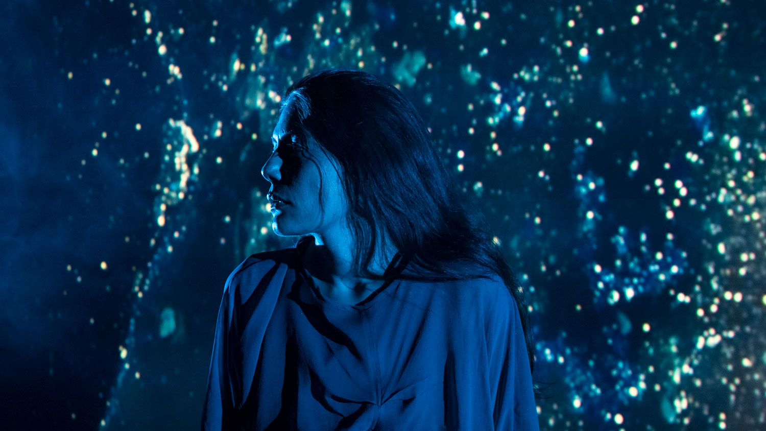 A female performer with long dark hair and wearing draped fabric looks over her shoulder in a room filled with blue starry light. 