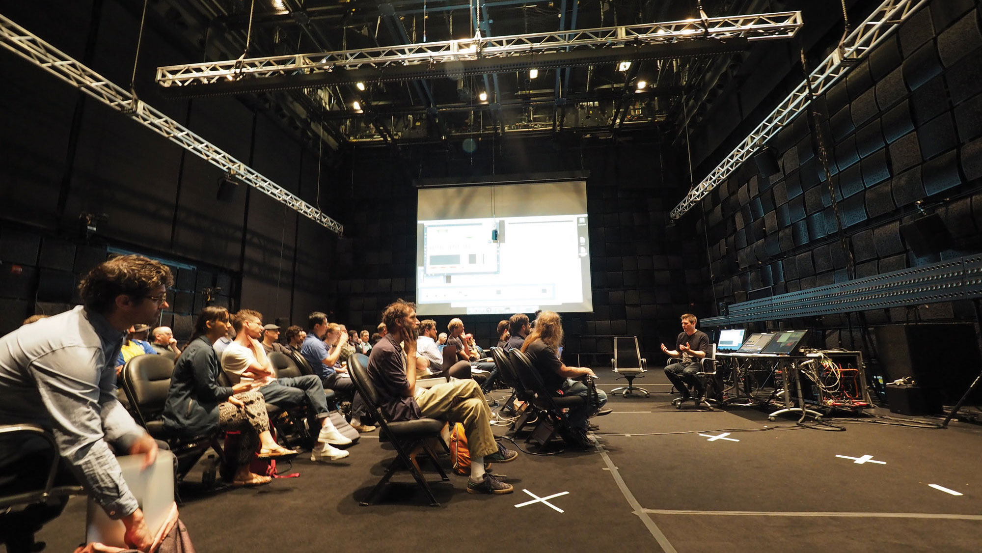 A crowd seated in a black box studio watching a man seated in the front of the room at a cluttered desk intently. 