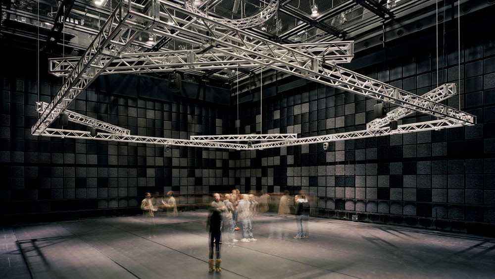A crowd blurred in motion under rigging suspended from the ceiling of a black box theater. 