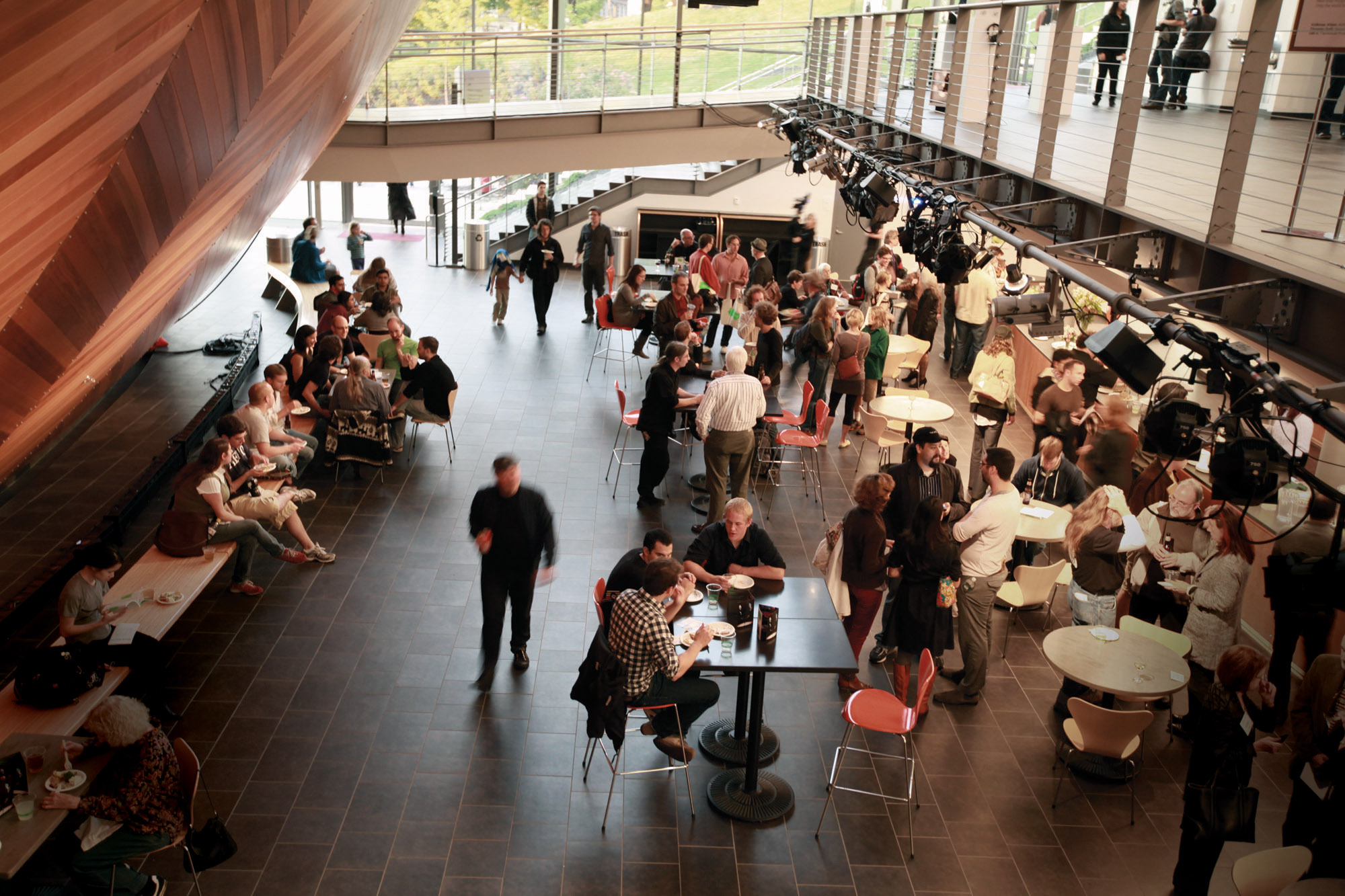 A crowd of people seated on on benches, at tables, and milling about in a cafe. 