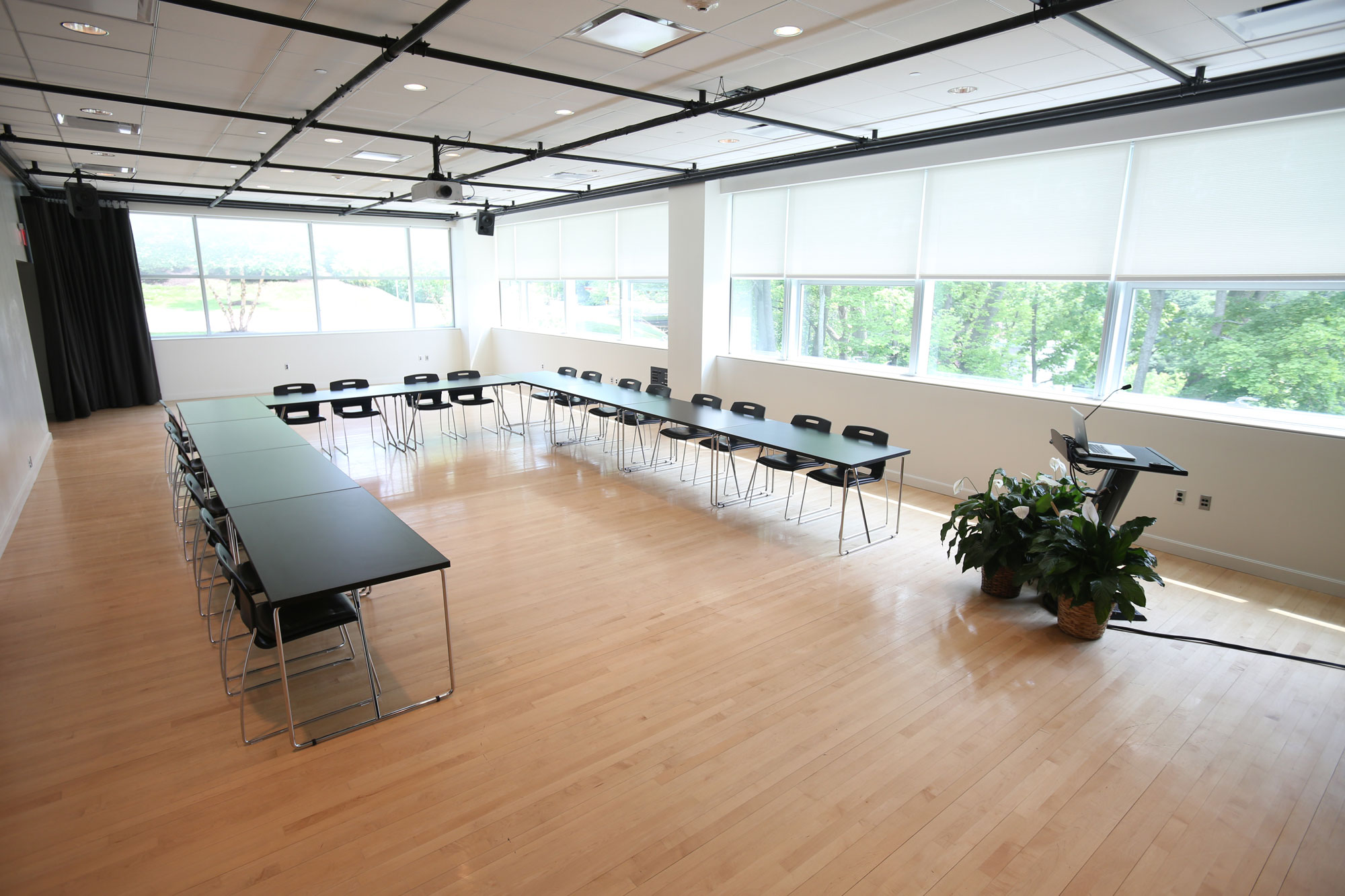 An empty studio with light wood floors, white walls, and black rigging on the ceiling flooded with natural light. A series of black tables are arranged in a U shape, a podium with flowers is positioned in the front of the room. 