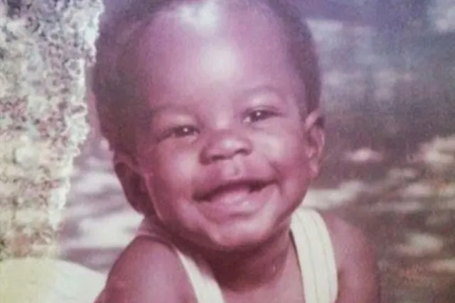 A Black baby wearing white overalls and posed in a studio portrait smiling contagiously. 
