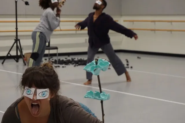 A woman with paper eyes taped over her eyes sticking out her tongue as dancers move through an empty dance studio in the background. 
