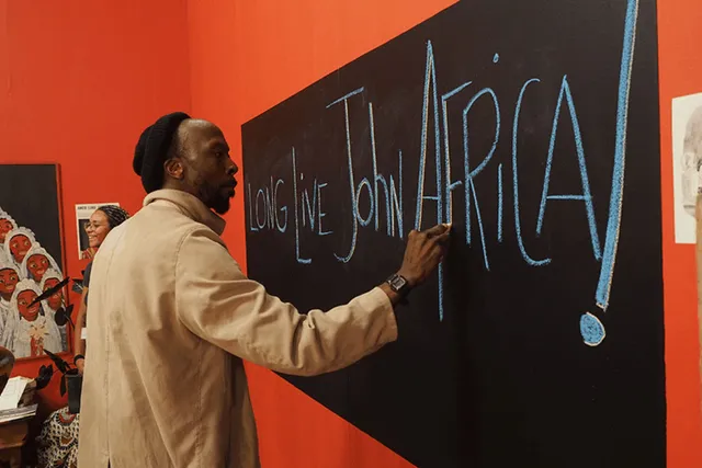 A Black man wearing a tan jacket and rolled black beanie writes on a blackboard "long live john africa!" in a red room. 