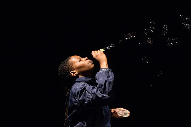 A Black person with long braided hair and wearing a blue shirt blows bubbles up into a dark room. 