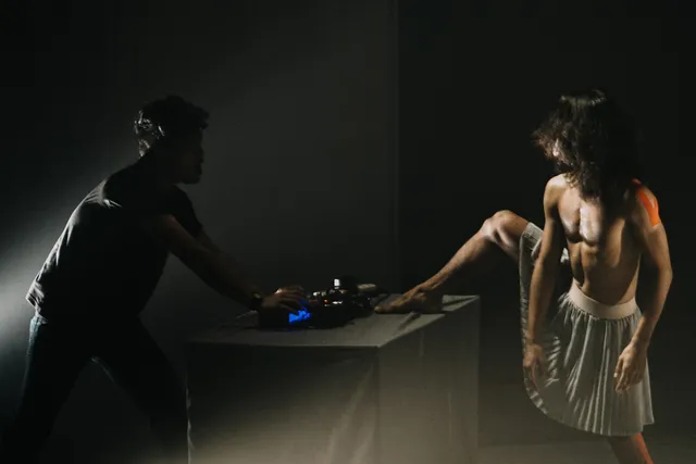 A shirtless person wearing a pleated skirt lifts their leg onto a keyboard behind controlled by a silhouetted figure on a black stage. 