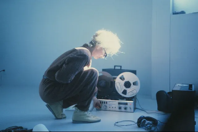 Maryanne Amacher kneeling in front of a 70's film projector in a cooly lit room.  