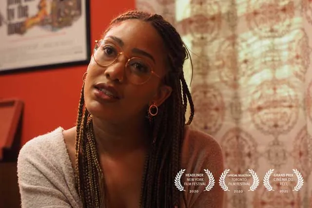 A Black woman wearing glasses and long braids looks toward the camera with head cocked in a room with red walls and patterned drapes. 