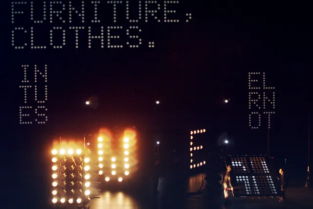 words spelled out by lights in a large black studio: furniture, clothes. in tu es.