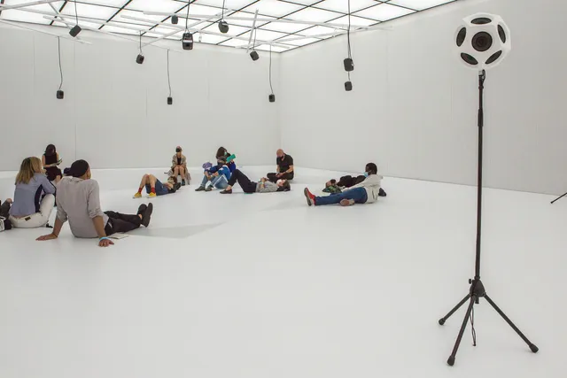 listeners sitting on the floor in a white gallery wtih speakers hung in a circle above