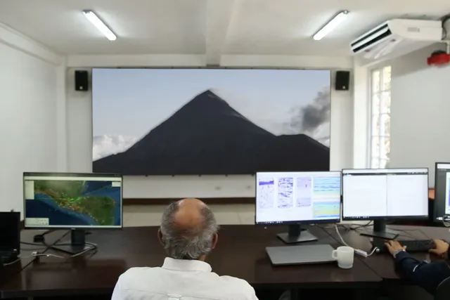 a man sitting in front of several computer screens looks at a large projection of a volcano