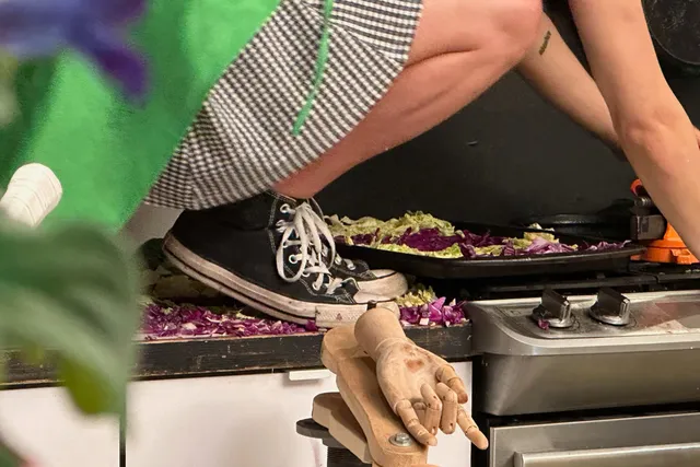A purple flower in a staged kitchen with one performer in black and white checkered costume and green apron on backwards crouching on top of a contemporary kitchen counter with hands working on the stovetop and chopped green and red cabbage strewn on all flat surfaces..