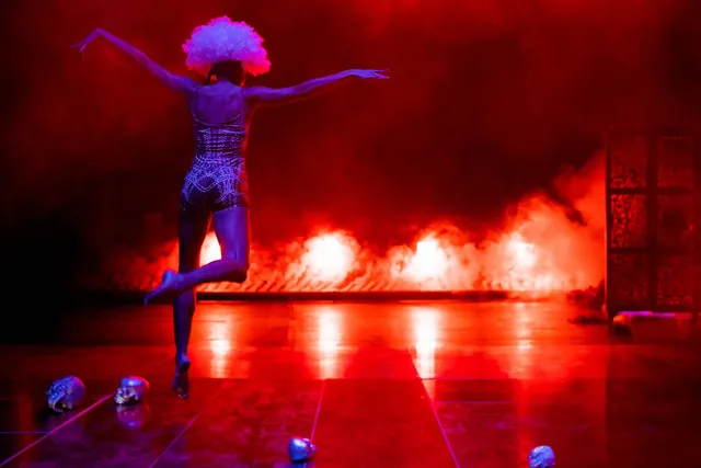 a woman runs toward the back of a stage her arms spread like an eagle, bright fire-like floor lights and fog in the background