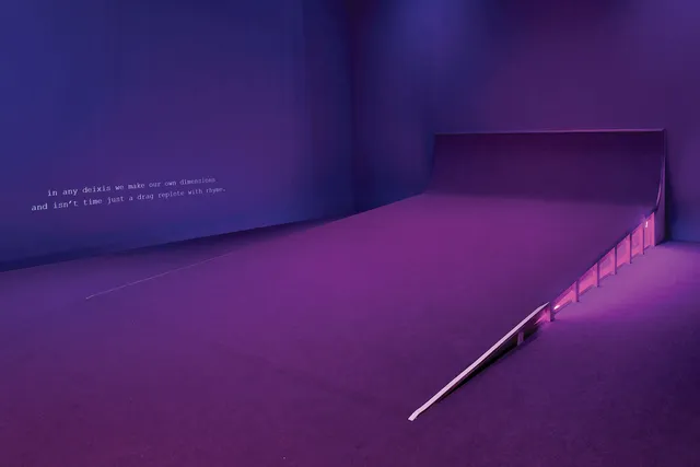 A large purple ramp with a slight incline in a room with purple walls and dim light. There is a caption low on the left wall and light shining from beneath the ramp.