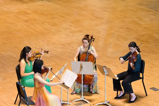 Four musicians performing in a string quartet setting on a wooden stage facing each other, two playing violins, one playing the cello, and one reading sheet music while playing the viola.