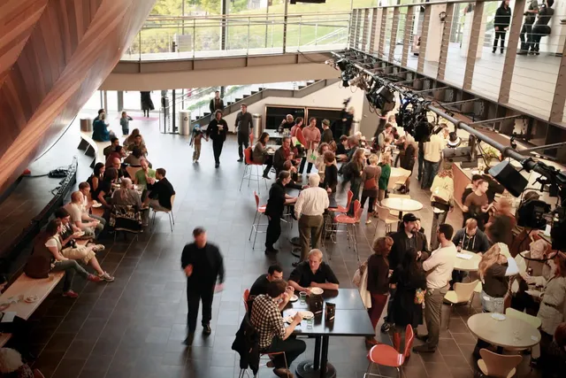 A crowd of people seated on on benches, at tables, and milling about in a cafe. 