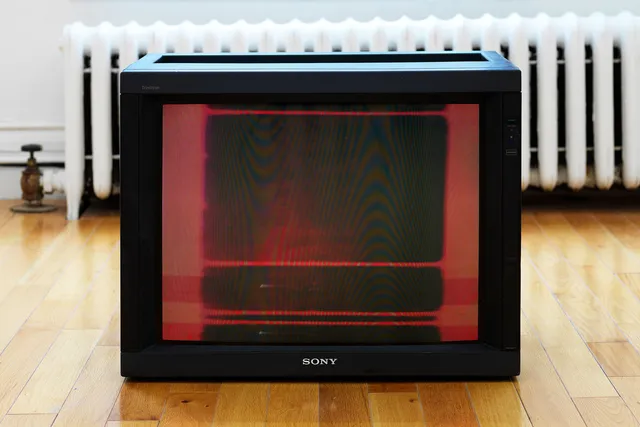 On a CRT monitor placed on a blonde gallery floor, a completely red frame from a video is visible on screen.