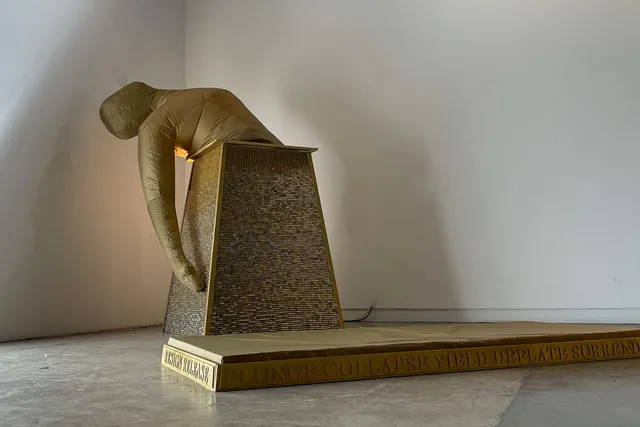 a deflated top half of a golden blowup body slumps over a golden pyramid shaped pedestal