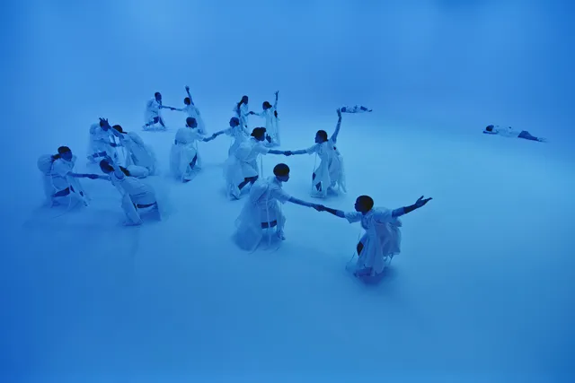 a group of dancers in white in a formation in a white cube bathed in blue light