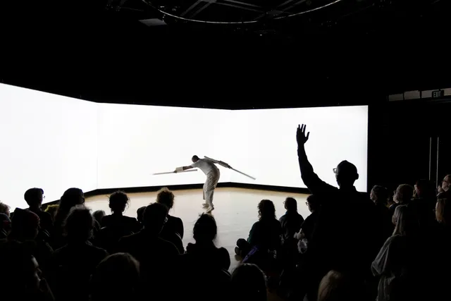 a black man in front of three large white screens in a black studio, audience in the foreground in darkness.