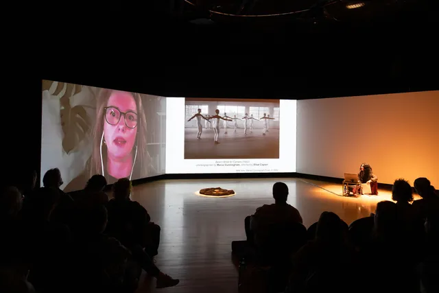three screens in a dark studio with audience in the foreground and a woman on a video conference on one screen in the background joined by a woman sitting in front of the right hand screen and images in the middle