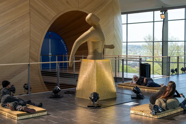 an inflatable sculpture of an amorphus human in a building lobby with people laying on platforms in the foreground