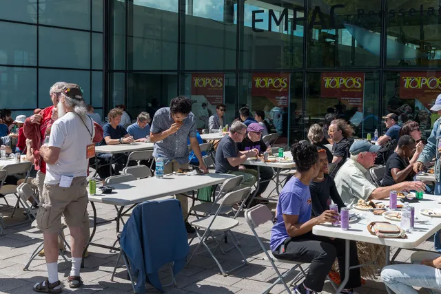 many people sitting at long tables in front of a glass building eating on a sunny day