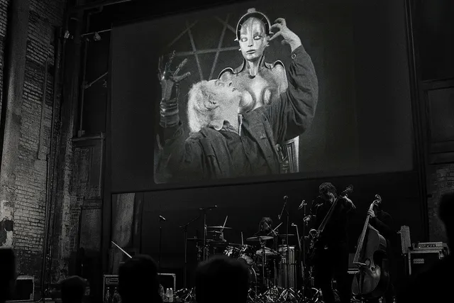 A black and white photograph of two musicians, [l-r] a guitarist and drummer, performing beneath a large screen on which a celebrated frame from Fritz Lang’s silent film Metropolis is projected.